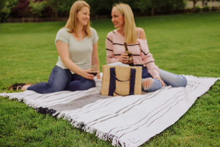 Two women enjoying a picnic on a blanket, with a basket of food beside them in a sunny outdoor setting.